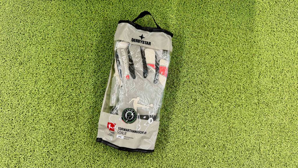 Torwarthandschuhe im Test: Ein Paar liegt in Originalverpackung auf einem Kunstrasen.