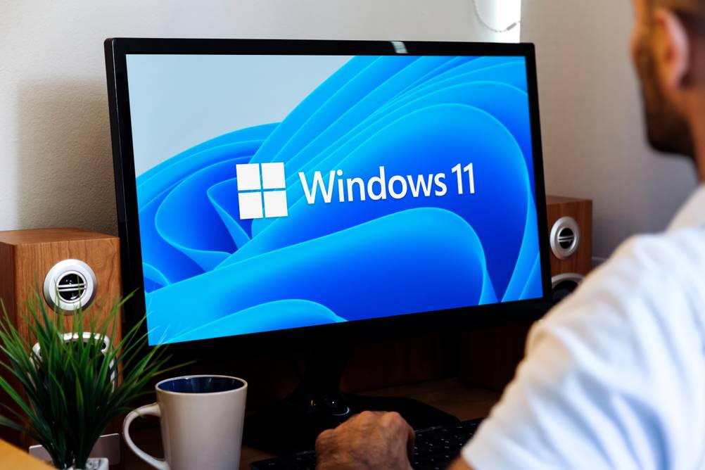 Desktop-PCs im Test: Ein Mann sitzt vor einem Monitor auf dem das Windows 11 Logo angezeigt wird.