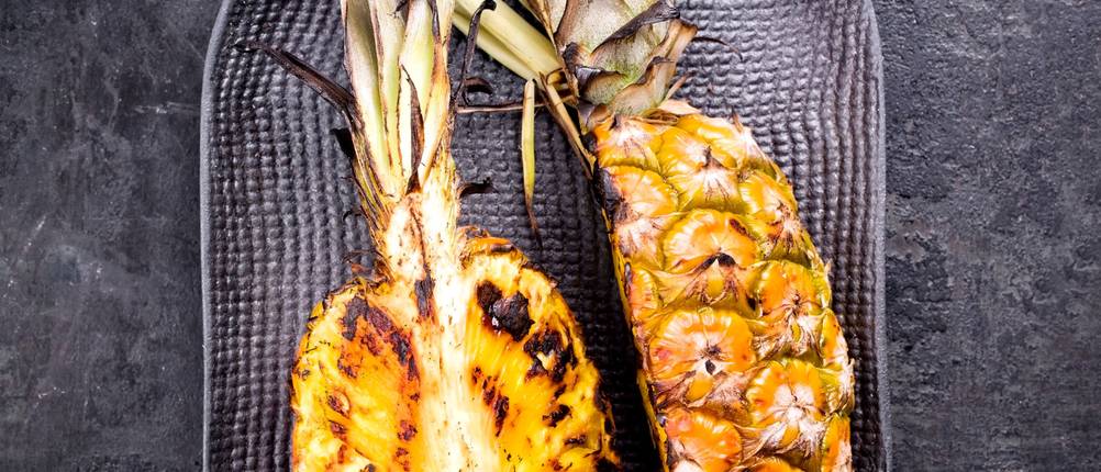 Gegrillte Ananas wird auf einer schwarzen Servierplatte auf dunklem Untergrund serviert.