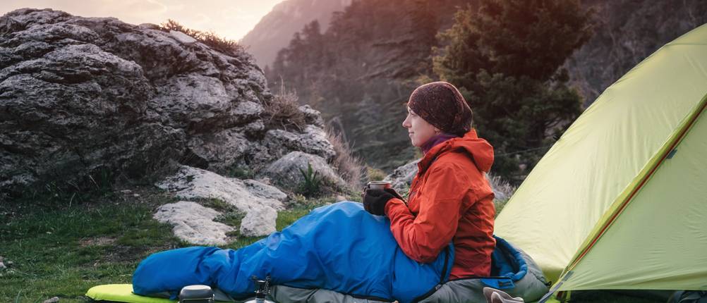 deuter-schlafsack-test: eine person liegt in einem schlafsack auf einer isomatte vor einem zelt. 