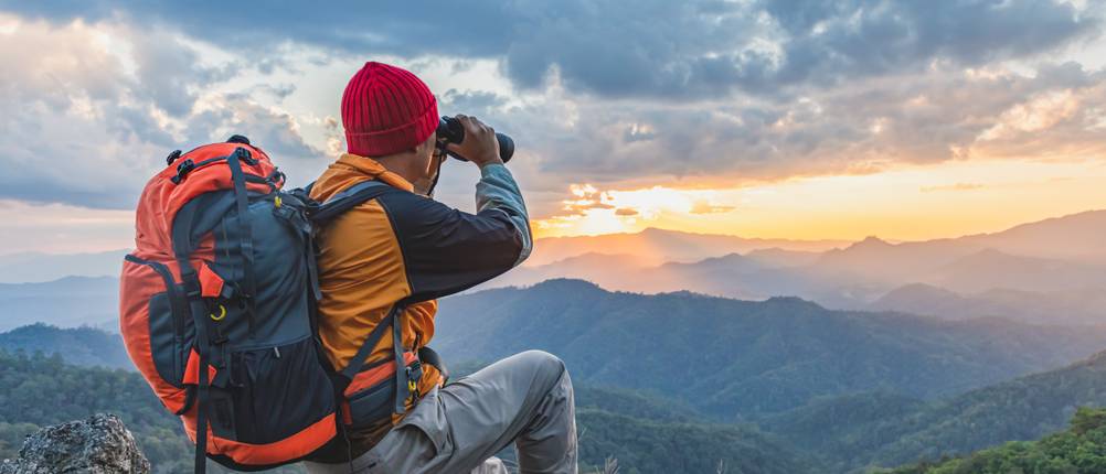 Deuter-Tagesrucksack-Test: Wanderer mit Rucksack und Fernglas in der Hand sitzt auf dem Gipfel eines Bergs und schaut in die Ferne.