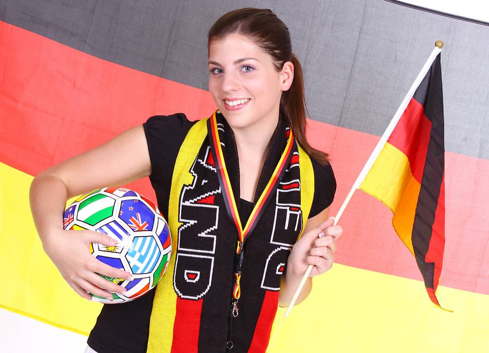 Deutschland-Schal-Test: Eine Frau mit Deutschland-Schal und Deutschland-Flagge mit einem Fußball in der Hand steht vor einer Deutschlandfahne.