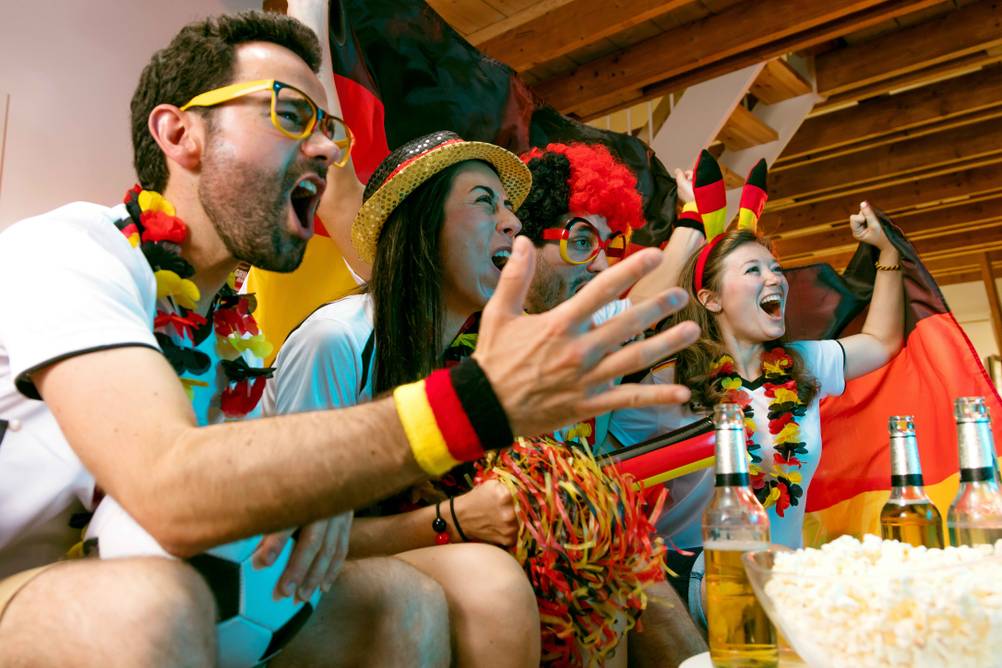 Deutschland-Fans mit Hüten und Trikots in einem großen Raum vor einer Leinwand oder einem Fernseher.
