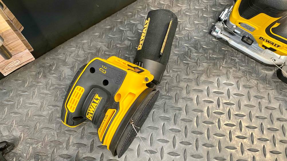 DeWalt Exzenterschleifer im Test: Ansicht auf ein gelb-schwarzes, liegendes Schleifgerät, inklusive Soft-Griff und Kletterverschlusssystem auf silberfarbener Metalloberfläche.