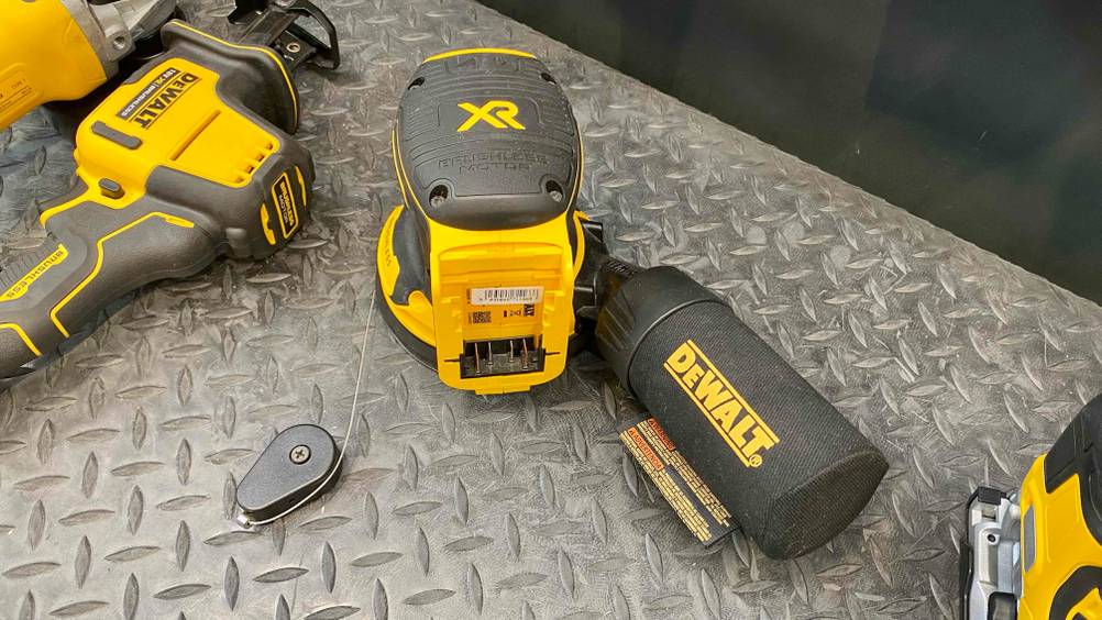 Exzenterschleifer-Test: Ansicht auf den Adapter für den Akku eines gelb-schwarzen Schleifgeräts der Marke DeWalt, inklusive Soft-Griff, ausgestellt mit anderen DeWalt Geräten auf silberfarbener Metallfläche.
