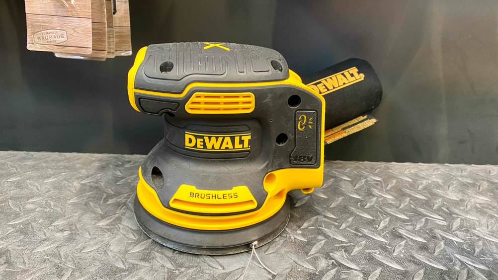 Exzenterschleifer im Test: Nahaufnahme eines gelb-schwarzen Schleifgeräts von DeWalt, inklusive Soft-Griff und Kletterverschlusssystem, das auf einer silberfarbener Ausstellungsfläche steht.
