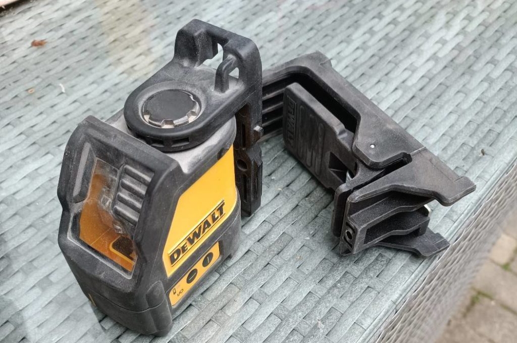 Batteriebetriebener Kreuzlinienlaser im Test: das Dewalt Modell befindet sich inklusive schwarzer Halterung auf einem hellgrauen Untergrund im Außenbereich.
