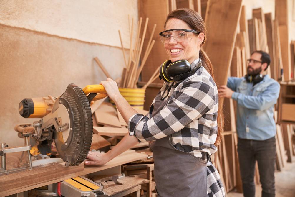 DeWalt-Kappsäge im Test: Eine junge Frau arbeitet an der Säge, während ein Mann im Hintergrund eine Holzbrett aus einem Regal nimmt.