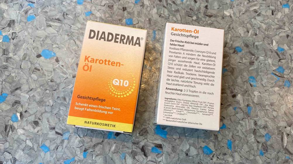 Diaderma Karottenöl im Test: zwei weiß-orange Verpackungen befinden sich nebeneinander auf einer blau-grau gesprenkelten Fläche, mit Blick auf die Vorder- sowie Rückseite inklusive Produktbeschreibung.