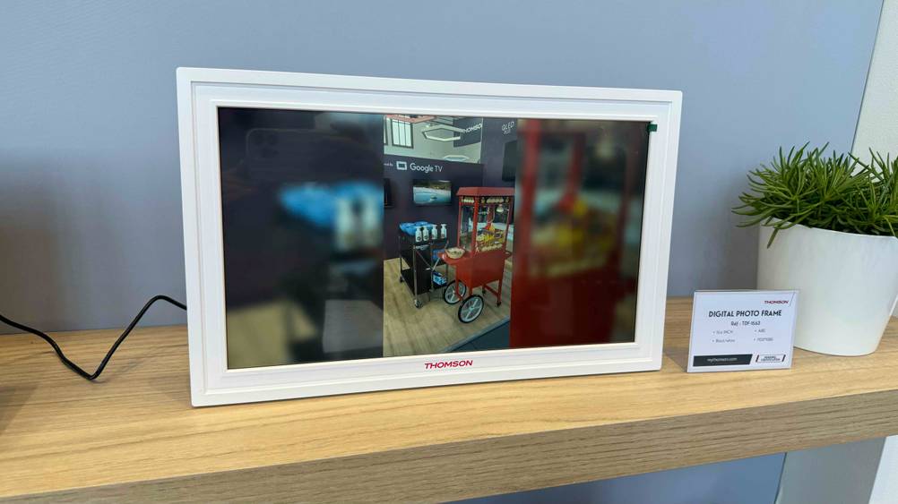 Ein getesteter Digitaler Bilderrahmen (15 Zoll) auf einem Holzregal mit Infotafel und Pflanze.