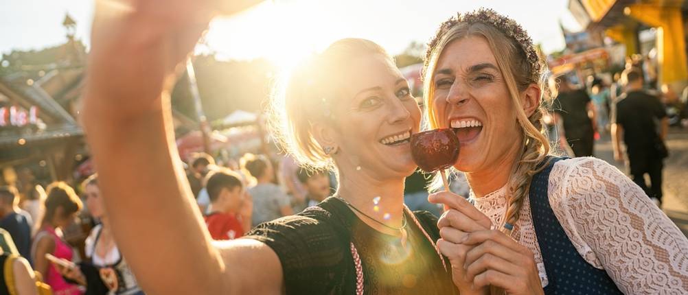 Dirndl Damen Test: Zwei Frauen lachen für ein Selfie in die Kamera und halten einen kandierten Apfel vor sich, im Hintergrund ist das Gelände der Wiesn zu sehen.