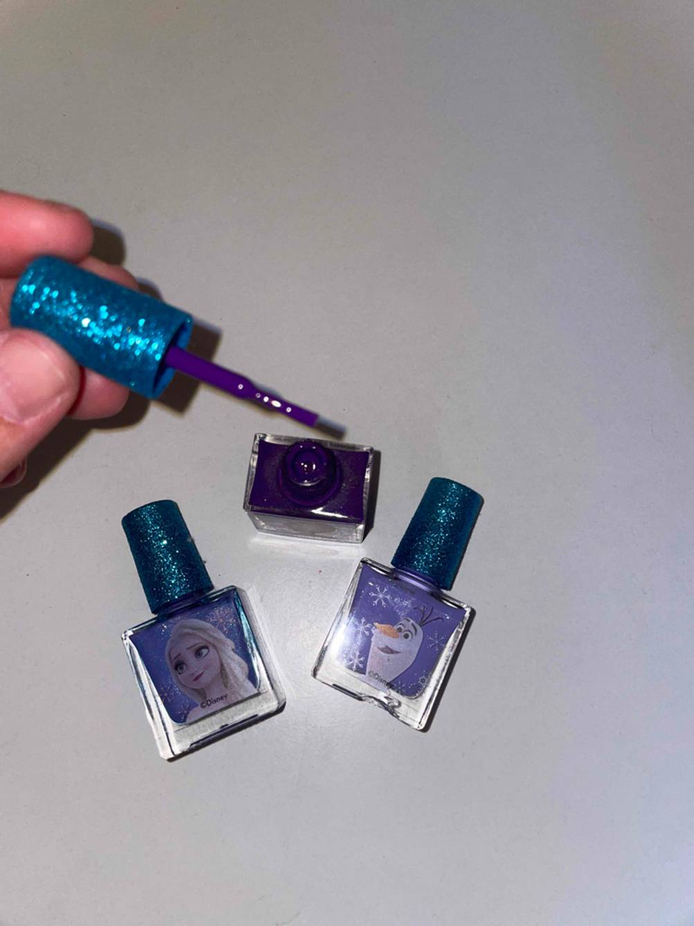 Im Test: Blick von oben in ein geöffnetes Fläschchen Nagellack für Kinder, eine Hand hält den Griff, zwei weitere Nagellacke liegen daneben.