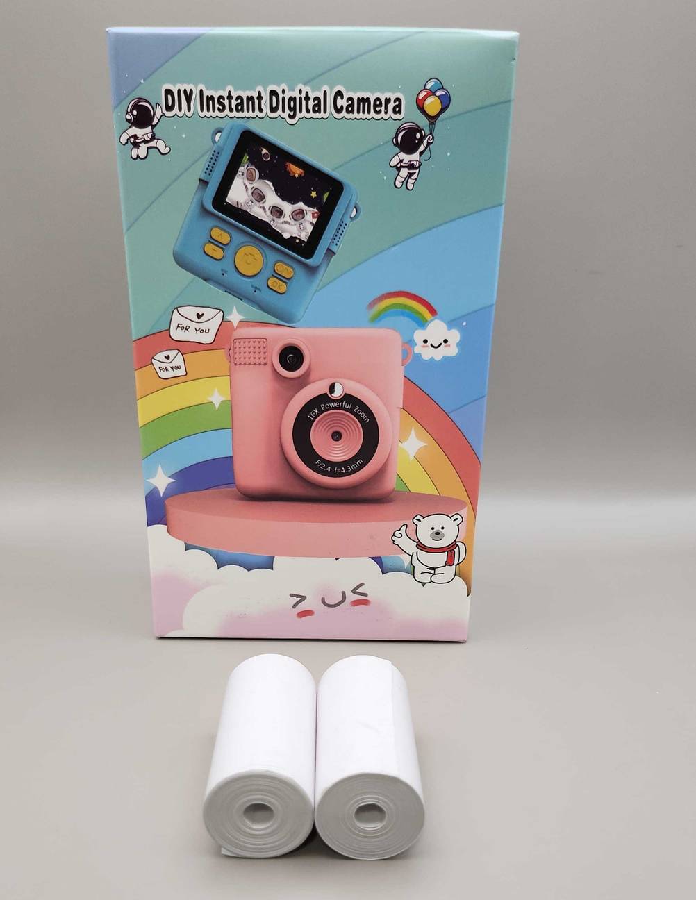 Getestet: Eine Kinderkamera in der Originalverpackung steht hinter zwei Rollen Fotopapier auf einer weißen Oberfläche.