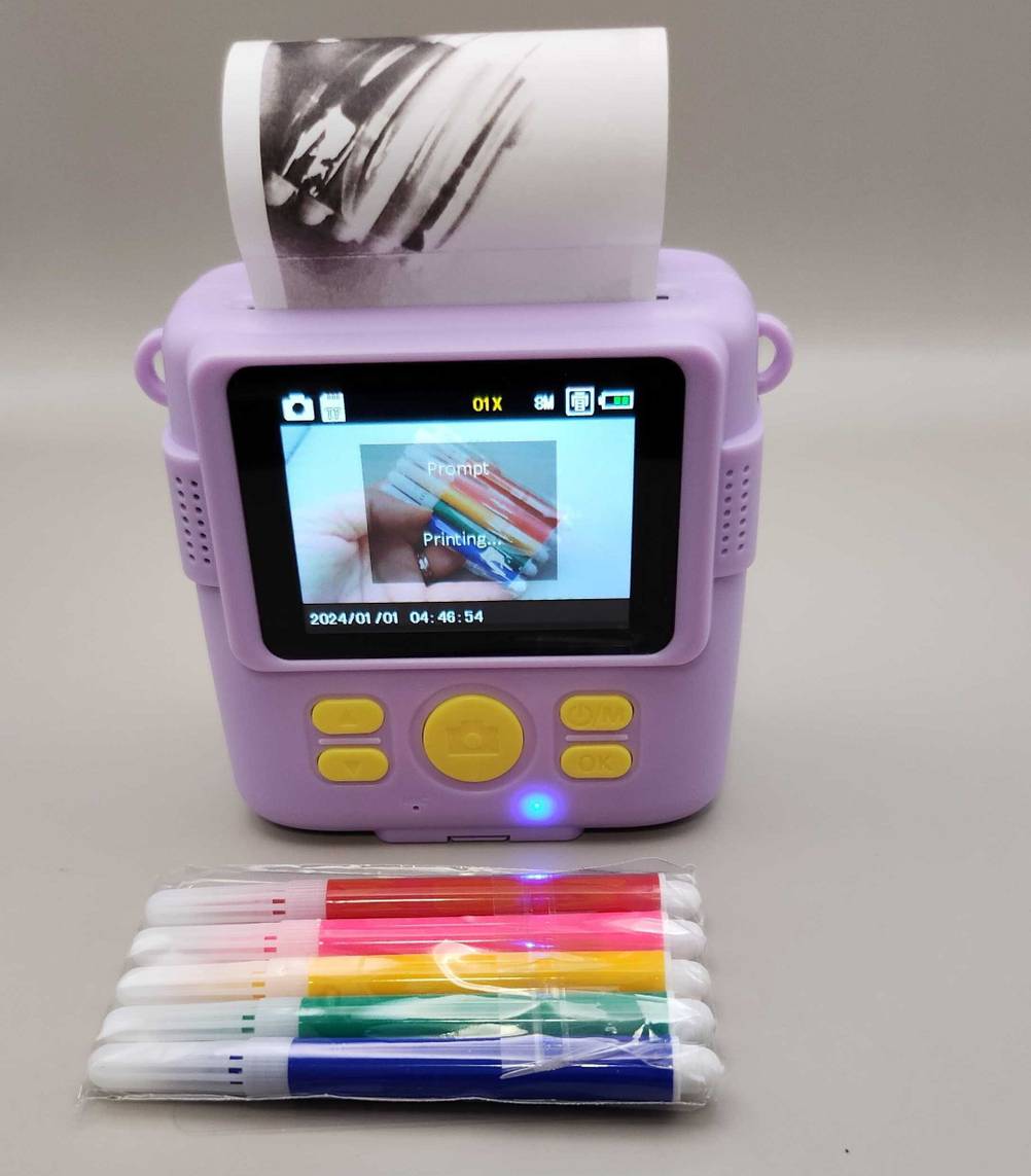 Eine getestete Kinderkamera in Lila inklusive Sofortbildauswurf steht hinter einer Packung Buntstifte auf einer weißen Fläche.