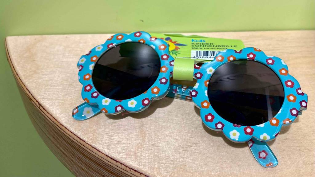 Babysonnenbrille-Test: Blaue Baby Sonnenbrille mit bunten Blümchenaufdruck und Etikett liegt auf einer Holzablage.