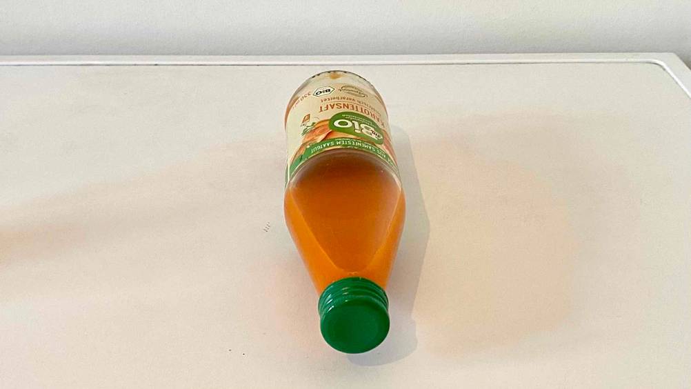 Eine Flasche getesteter Karottensaft liegt mit dem Deckel nach vorn auf weißer Fläche.
