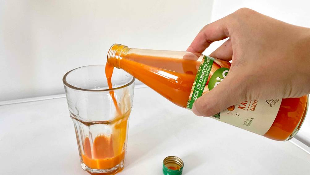 Karottensaft im Test: Hand schenkt aus einer geöffneten Flasche in ein Glas ein.