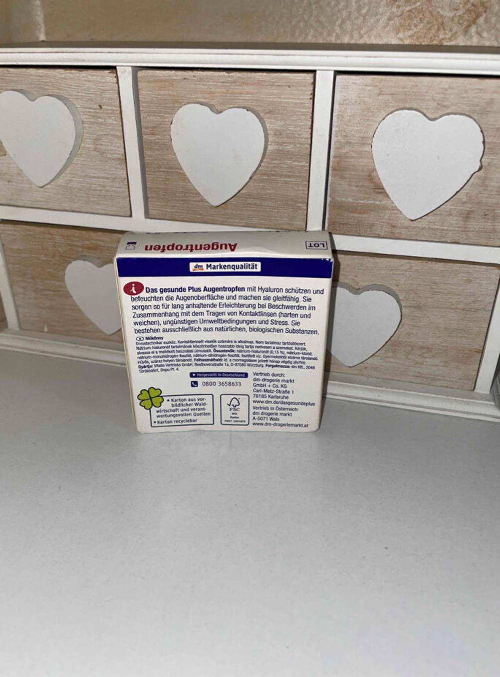 Eine Packung der getesteten Augentropfen steht vor einer kleinen Holzbox mit der Rückseite nach vorne.