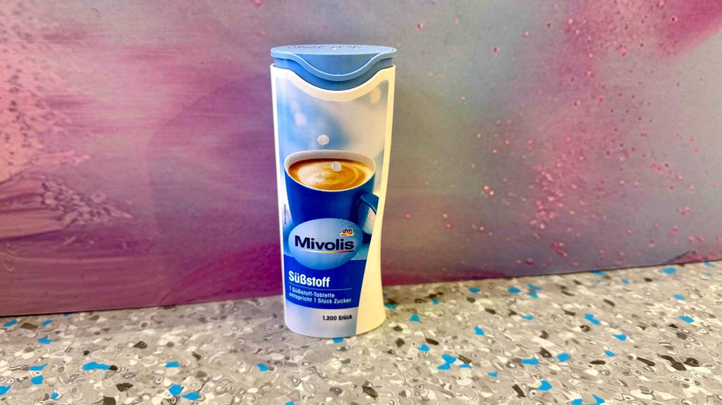 Ein Dosenspender mit getesteten Süßstoff-Tabletten steht vor einer lila Wand auf einer gesprenkelten Fliese.