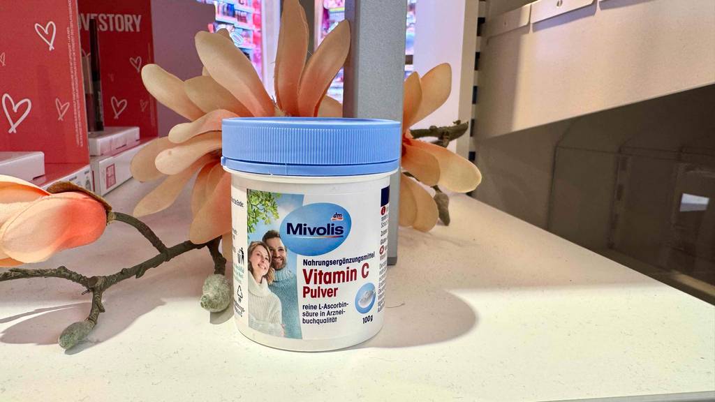 Eine Dose Vitamin-C-Pulver steht zum testen vor einer Blume auf einem weißen Regalbrett.