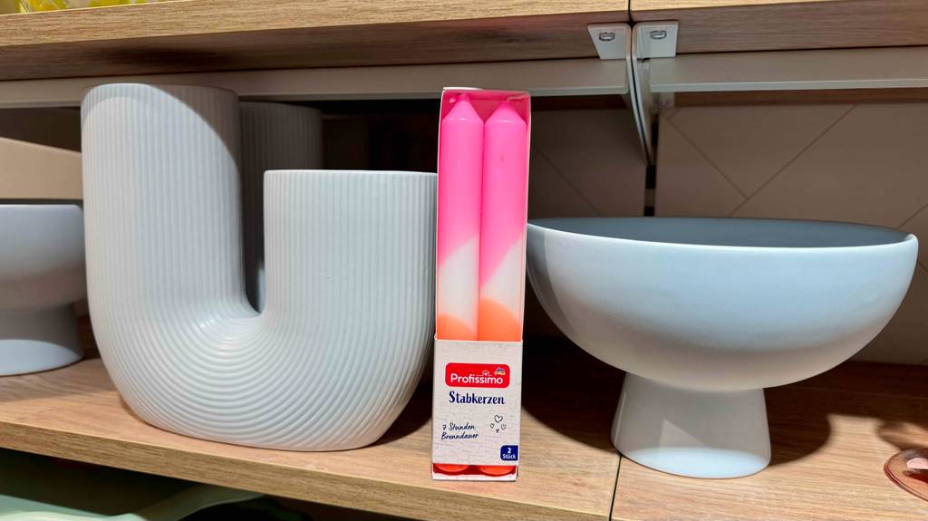 Im Test: Eine 2er-Packung mit rosa-weiß-orangen Stabkerzen steht vor Keramikformen in einem Holzregal.