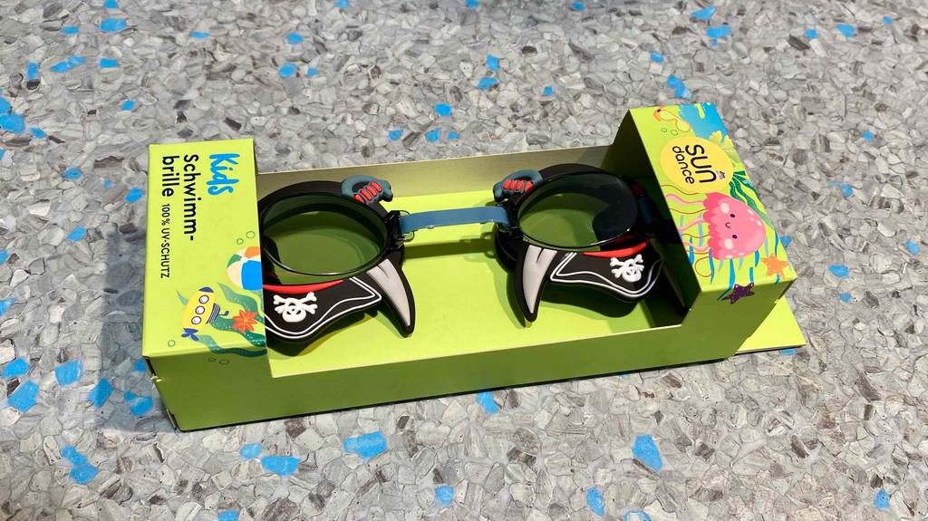 Schwimmbrille-Test: Eine Sun Dance Piraten-Schwimmbrille im Test liegt in ihrer hellgrünen Verpackung auf einem blau-grau gesprenkelten Bodenbelag.