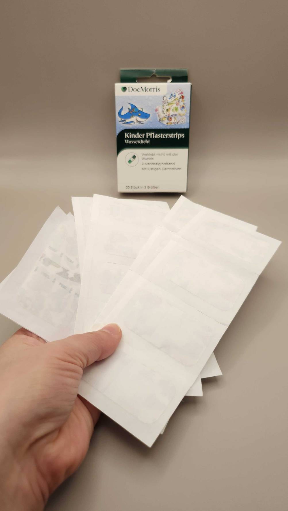 Eine Hand hält mehrere Pflasterstreifen über einer beigefarbenen Fläche, auf der eine Verpackung mit Kinder-Pflasterstrips steht.