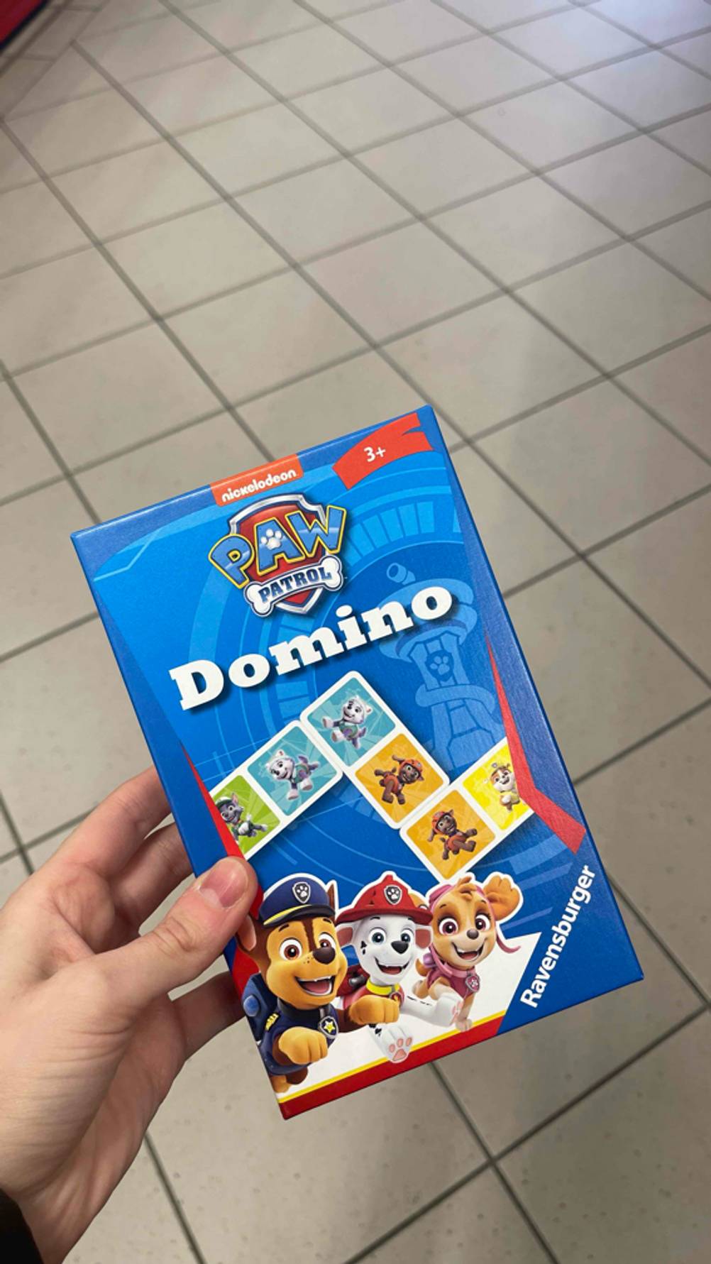 Eine Hand hält die blaue Ravensburger-Packung mit dem getesteten Domino-Spiel über einem weißen Fliesenboden.