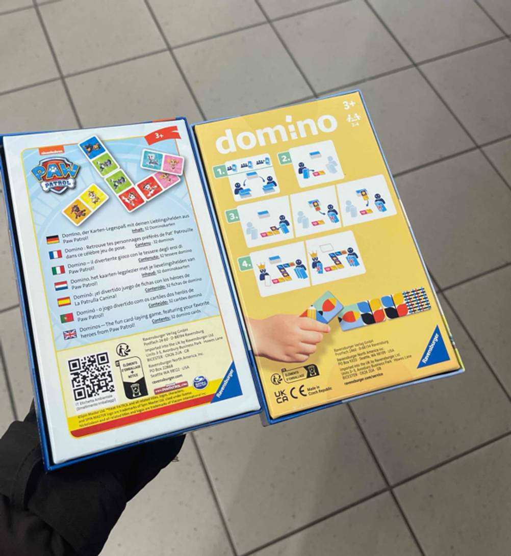 Eine Hand zeigt die beiden Spieleanleitungen auf den Rückseiten der Verpackungen mit den beiden Domino-Spielen im Test.