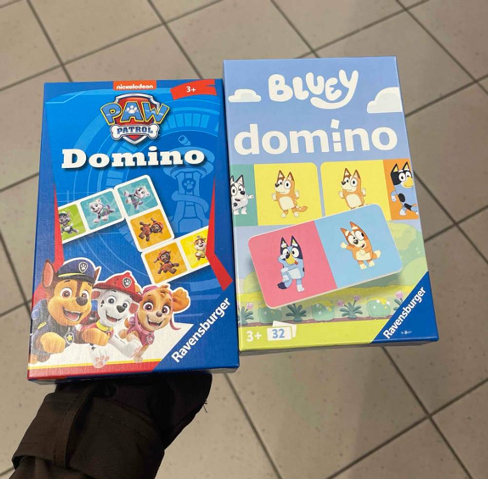 Eine Hand hält zwei unterschiedliche, getestete Domino-Spiele in blauen Verpackungen von Ravensburger über einem grauen Fliesenboden.