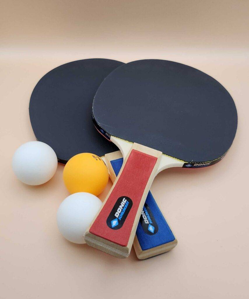 Getestetet: Zwei Tischtennisschläger liegen neben drei Tischtennisbällen auf einem beigefarbenen Untergrund.