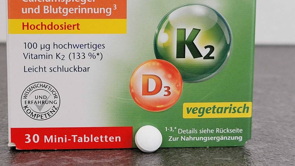 Eine Tablette des getesteten Vitamin K2 lehnt an ihrer Packung auf einer dunklen Oberfläche.