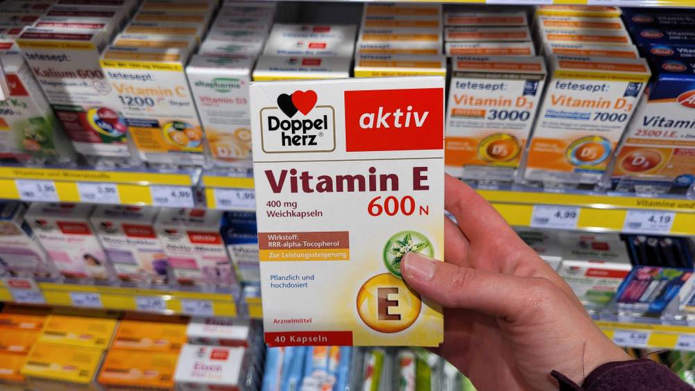 Eine Hand hält eine Packung Vitamin E vor ein Verkaufsregal mit vielen anderen Produkten.