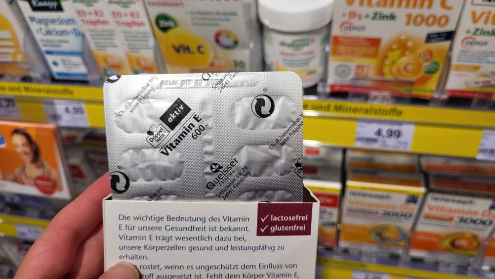 Eine Hand hält die Rückseite einer Packung getesteten Vitamin E vor ein Regal, bei der auch ein Tabettenblister mit der Rückseite hervor schaut.