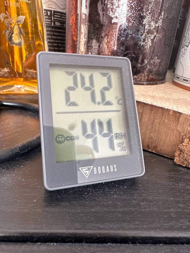 Viereckiges Doqaus Thermometer im Test steht mit eingeschaltetem Display auf einer schwarzen Fläche vor Dekogegenständen. hellem Hintergrund.