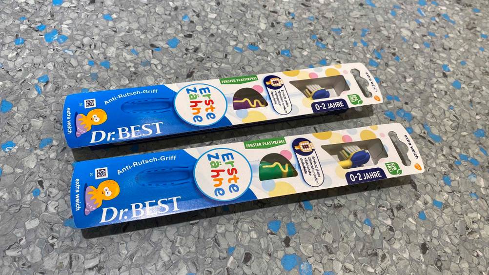 Baby-Zahnbürste im Test: zwei verpackte Modelle von Dr. Best liegt in ihren Originalverpackungen auf einem blau-grauen Boden.