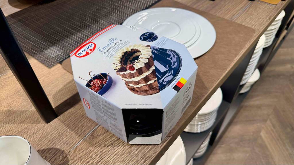 Backformen aus Emaille im Test: Eine Verpackung von Dr. Oetker inklusive schwarzem 18-cm-Gugelhupf liegt auf einem Regal mit weißen Tellern.