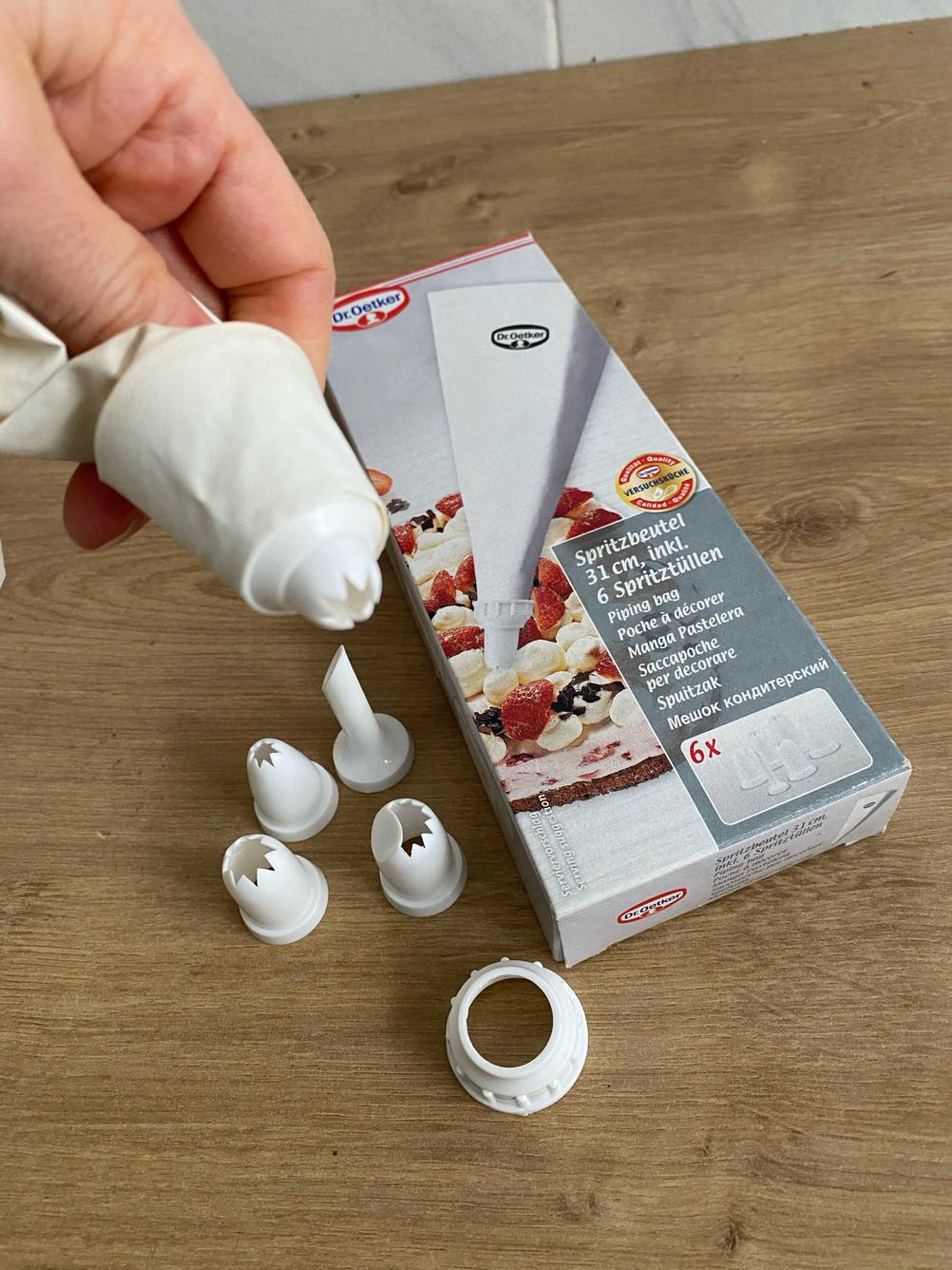 Im Test: Spritzbeutel aus hellem Gewebe von Dr. Oetker in einer Hand über verschiedenen Spritzaufsätzen und der Verpackung auf Holzuntergrund.