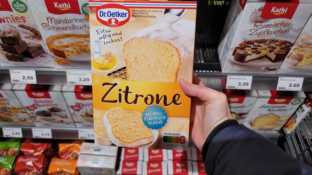 Eine Hand hält eine Packung Kuchen-Backmischung zum testen vor einem Verkaufsregal.