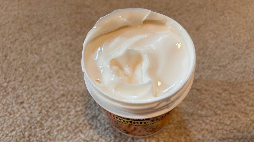 Schrundensalben-Test: ein geöffneter Tiegel mit cremiger Salbe auf einem beigefarbenen Teppichboden von oben betrachtet.