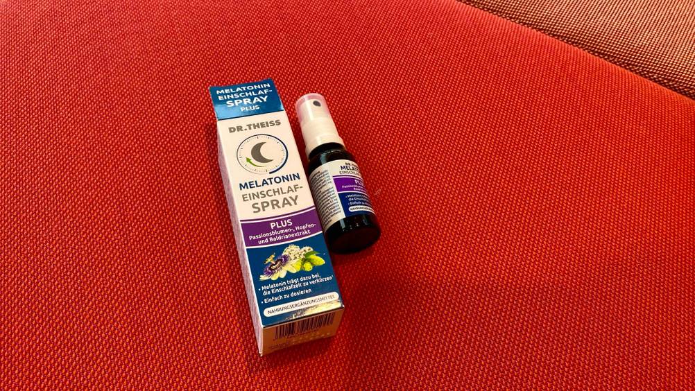 Ein getestetes Melatonin-Spray neben seiner Originalverpackung von schräg unten auf einer roten Fläche.