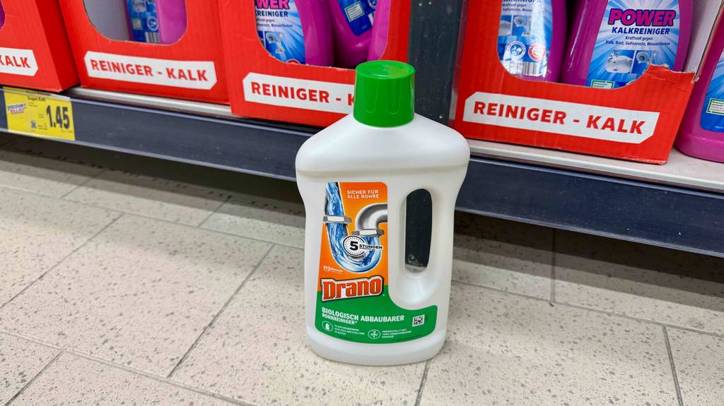 Abflussreiniger im Test: Eine weiße Flasche aus Kunststoff steht vor einem Regal mit Kalk-Reinigern auf dem Boden.
