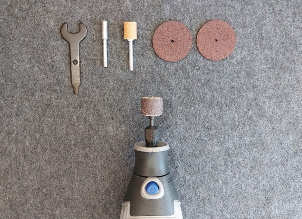 Ein Dremel 3000 Multifunktionswerkzeug liegt unterhalb mehrerer Zubehörteile, darunter Schleifaufsätze, Träger und ein Spannschlüssel. Alle Elemente sind auf grauem Untergrund ausgebreitet dargestellt.