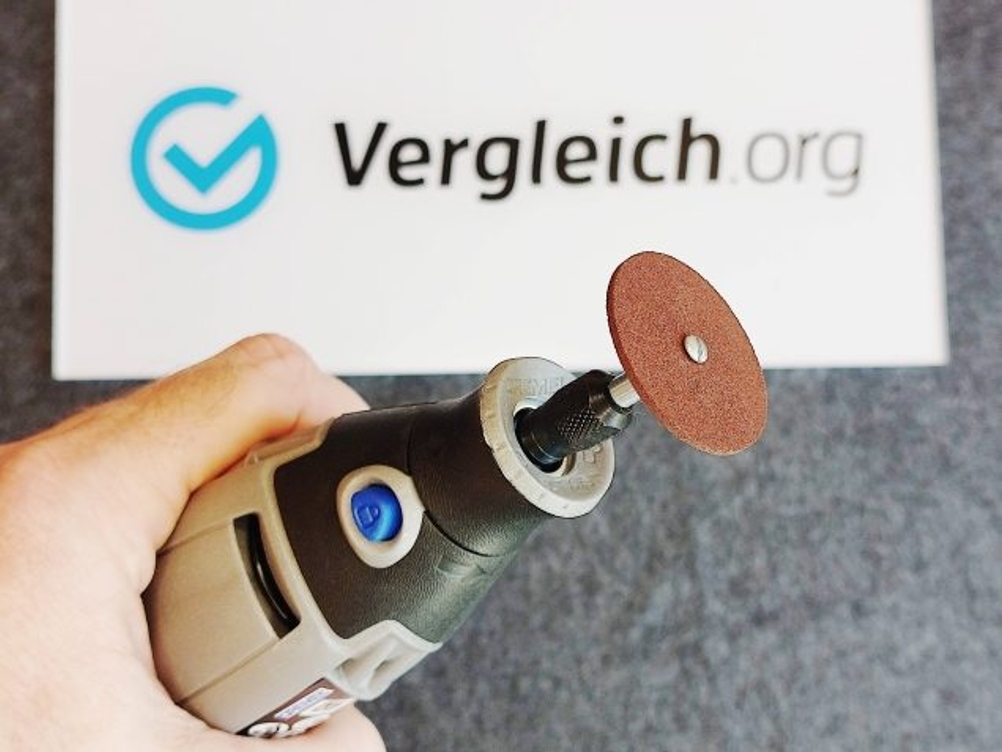 Ein Dremel-Multifunktionswerkzeug mit eingesetzter Schleifscheibe wird von einer Hand gehalten. Das Werkzeug ist startbereit und auf einen grauen Untergrund gerichtet.