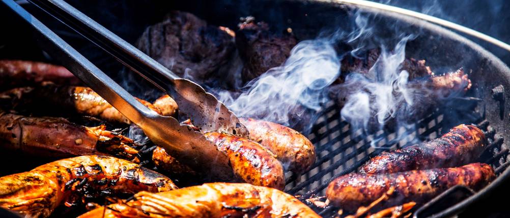 Auf einem rauchenden Grill mit diversem Grillfleisch wird eine Bratwurst mit einer Grillzange umgedreht.