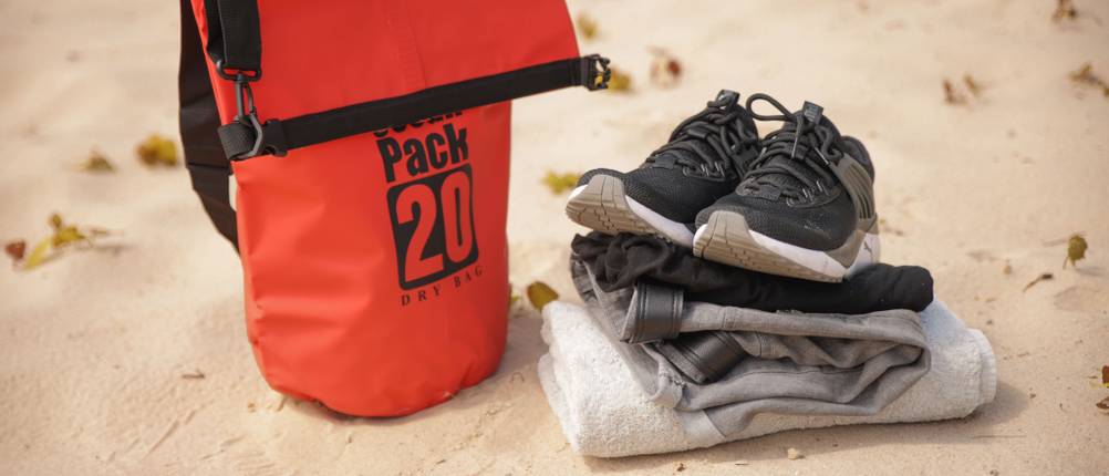 Dry-Bags im Test: Neben einem Dry-Bag liegen ein Handtuch, eine Hose, ein T-Shirt und Schuhe im Sand.