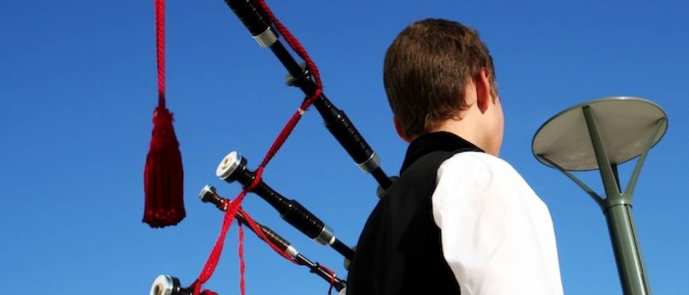 Dudelsack im Test. Ein junger Mann mit einem Dudelsack von hinten aufgenommen vor blauem Himmel.