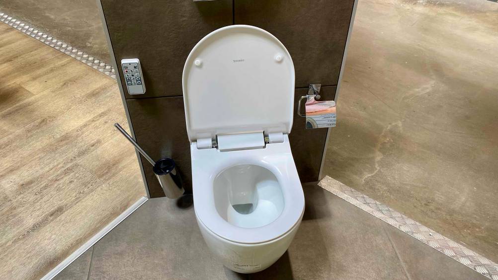 Ein Dusch-WC im Test von vorne mit geöffnetem Deckel an einer dunklen Wand montiert.