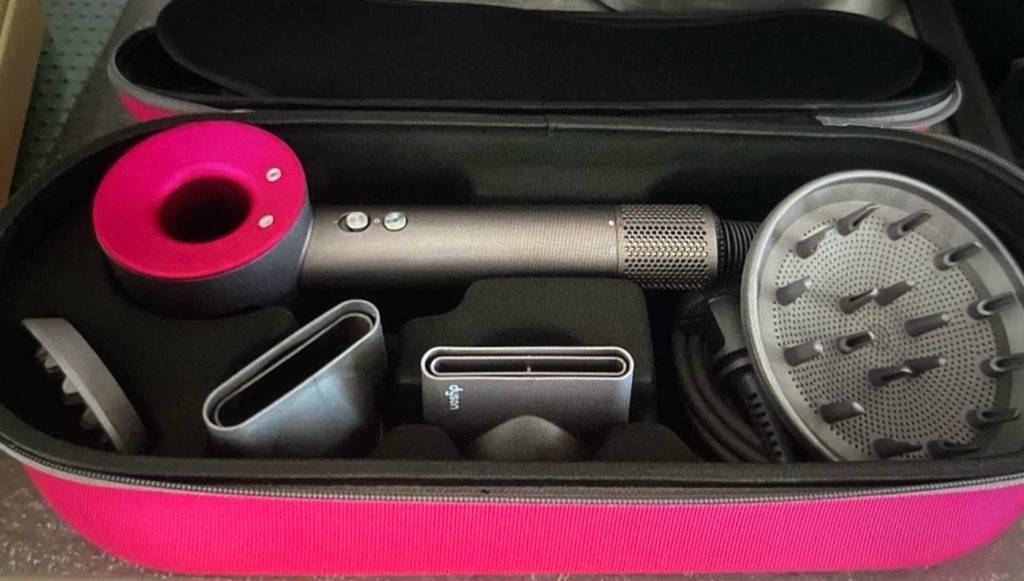 Haartrockner-Test: Nahaufnahme eines Dyson Modells im rosa-silbernen Design inklusive verschiedener Aufsätze in seiner geöffneten Aufbewahrungstasche mit Reißverschluss.