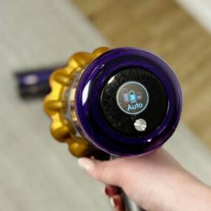 Das Display des Dyson V15 Detect zeigt den aktivierten Automatikmodus. Eine Hand hält das Gerät über einem hellen Teppichboden.
